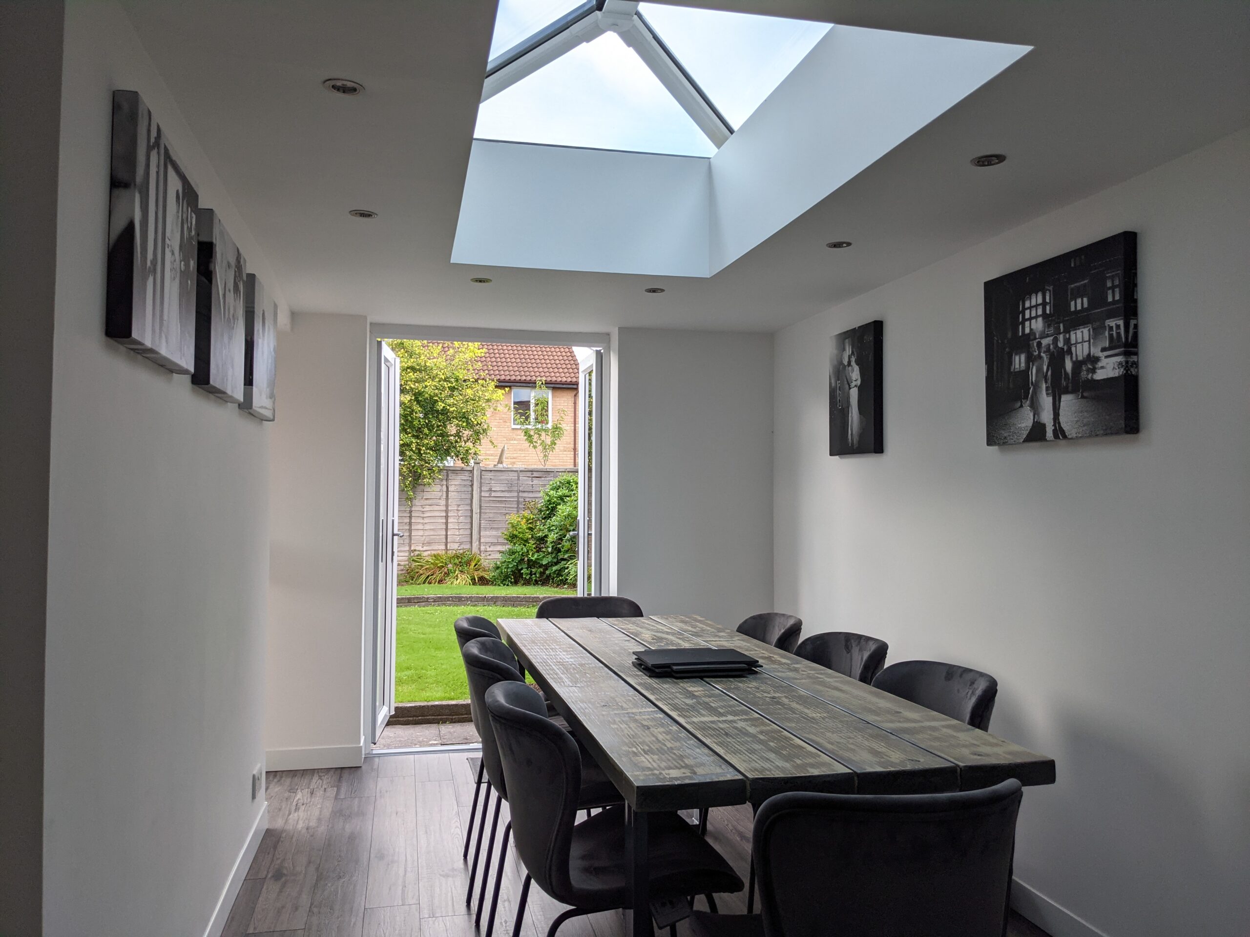 Minimalist dining room with a long table, skylight roof lantern, and open doors leading to a garden.
