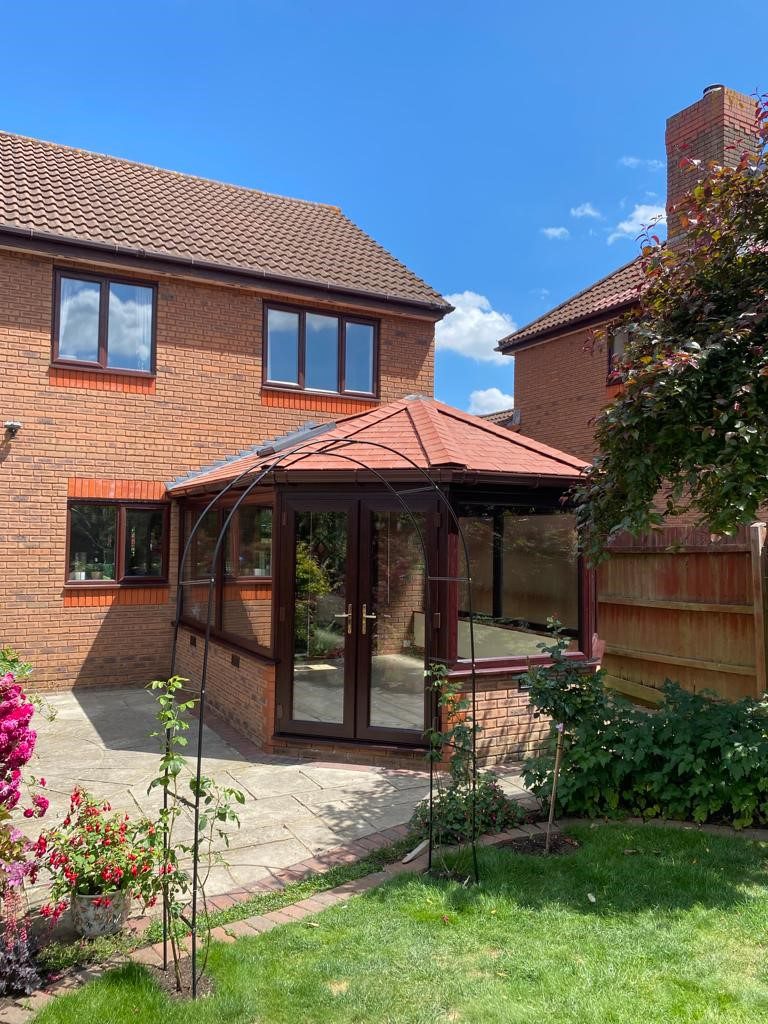 Rear view of a two-story brick house with a newly upgraded conservatory featuring a solid tiled roof in a matching red tone. The structure has large, dark-framed glass doors and windows, blending seamlessly with the house. 