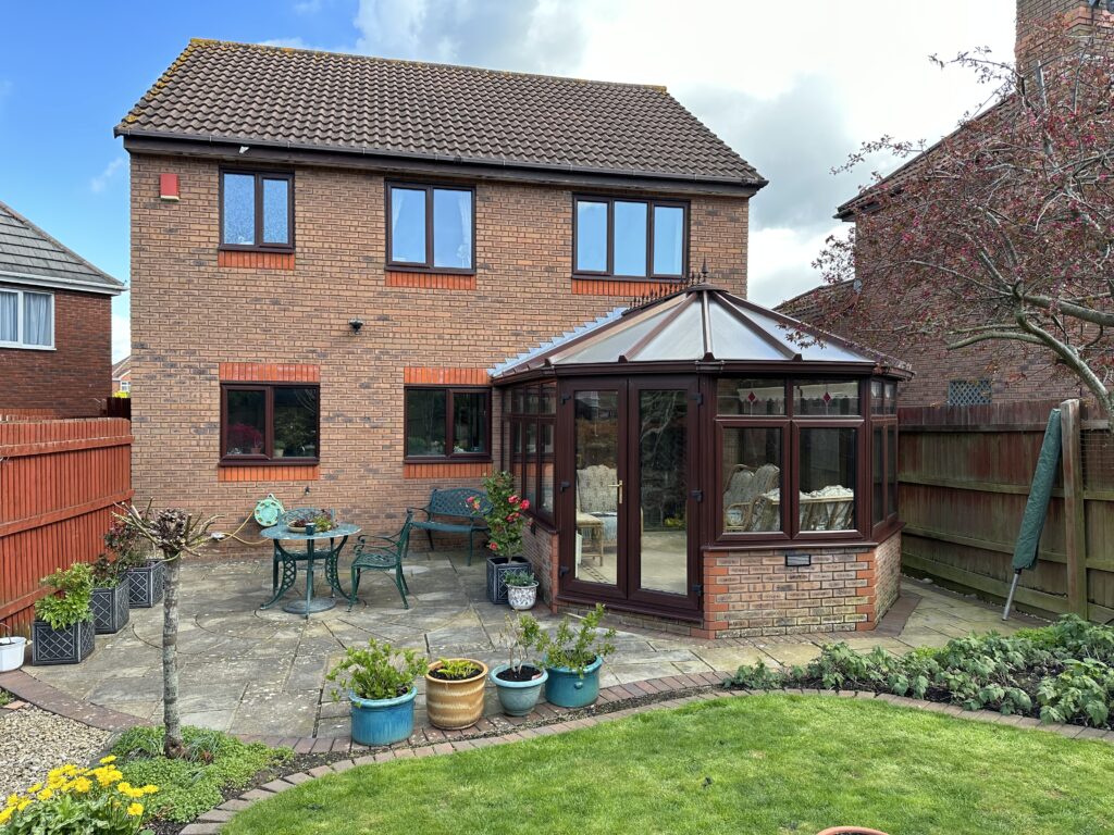 Rear view of a two-story brick house with an older-style conservatory featuring a translucent polycarbonate or glass roof. The conservatory has dark brown framing with clear windows and is furnished inside.