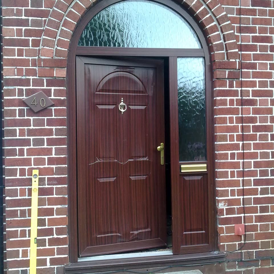 Old brown woodgrain-effect front door with semi-arched toplight and brick surround, slightly open showing brass handle and door knocker