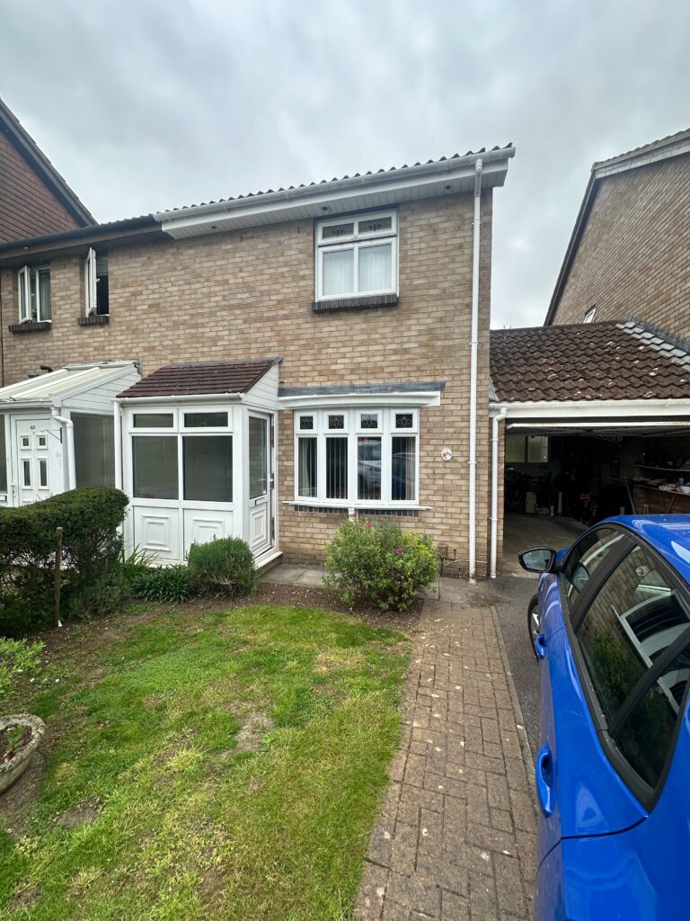 Front of a semi-detached house with an old white UPVC porch featuring a sloped roof. The garden has patchy grass and shrubs.
